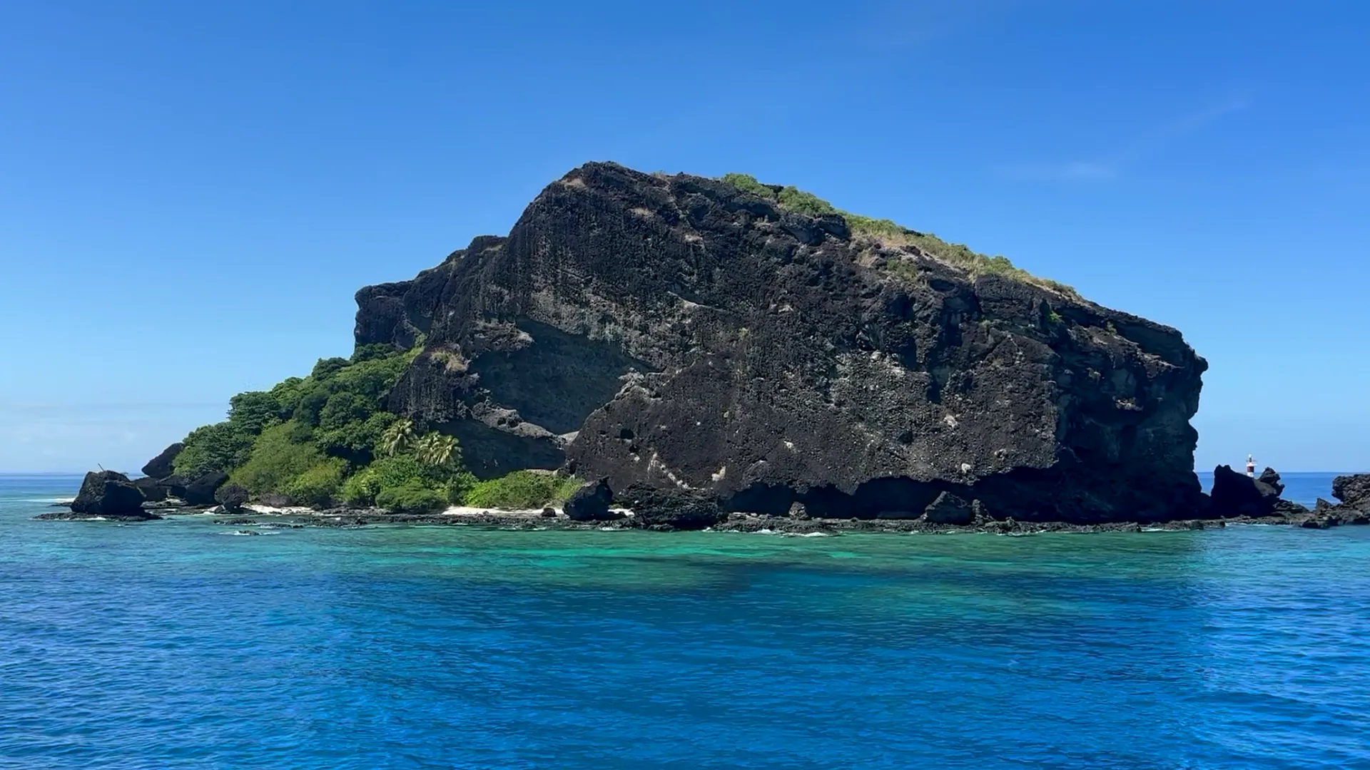 A dark rocky island with green vegetation and a turquoise reef, filmed from a boat off Fiji