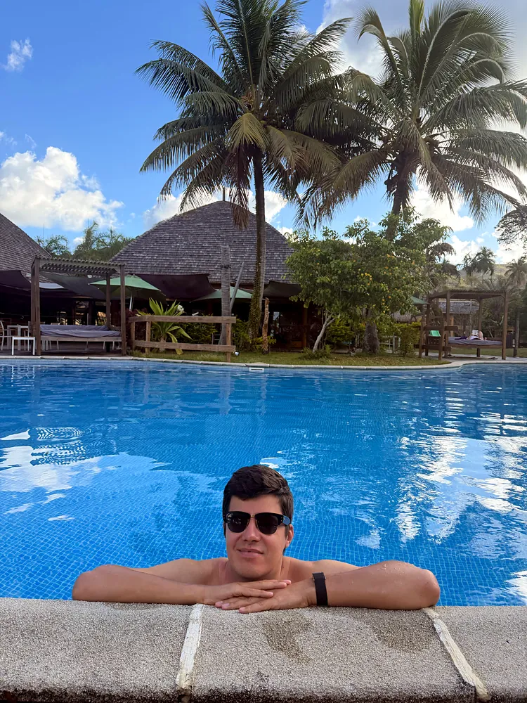 A man in sunglasses at the edge of a pool, with palm trees and a thatched pavilion behind