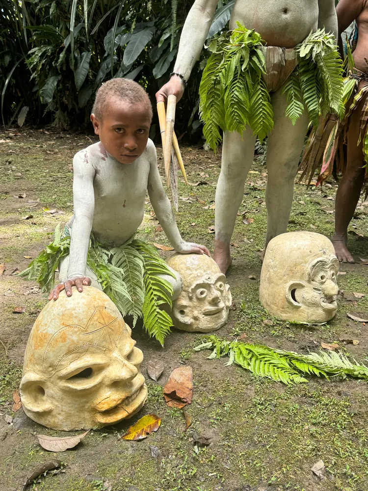 Мальчик с покрытым глиной телом и в юбке из папоротника рядом с тремя глиняными масками; за ним взрослые участники