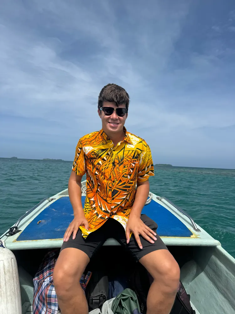 A young man in sunglasses and a patterned shirt sits on the bow of a boat in a lagoon