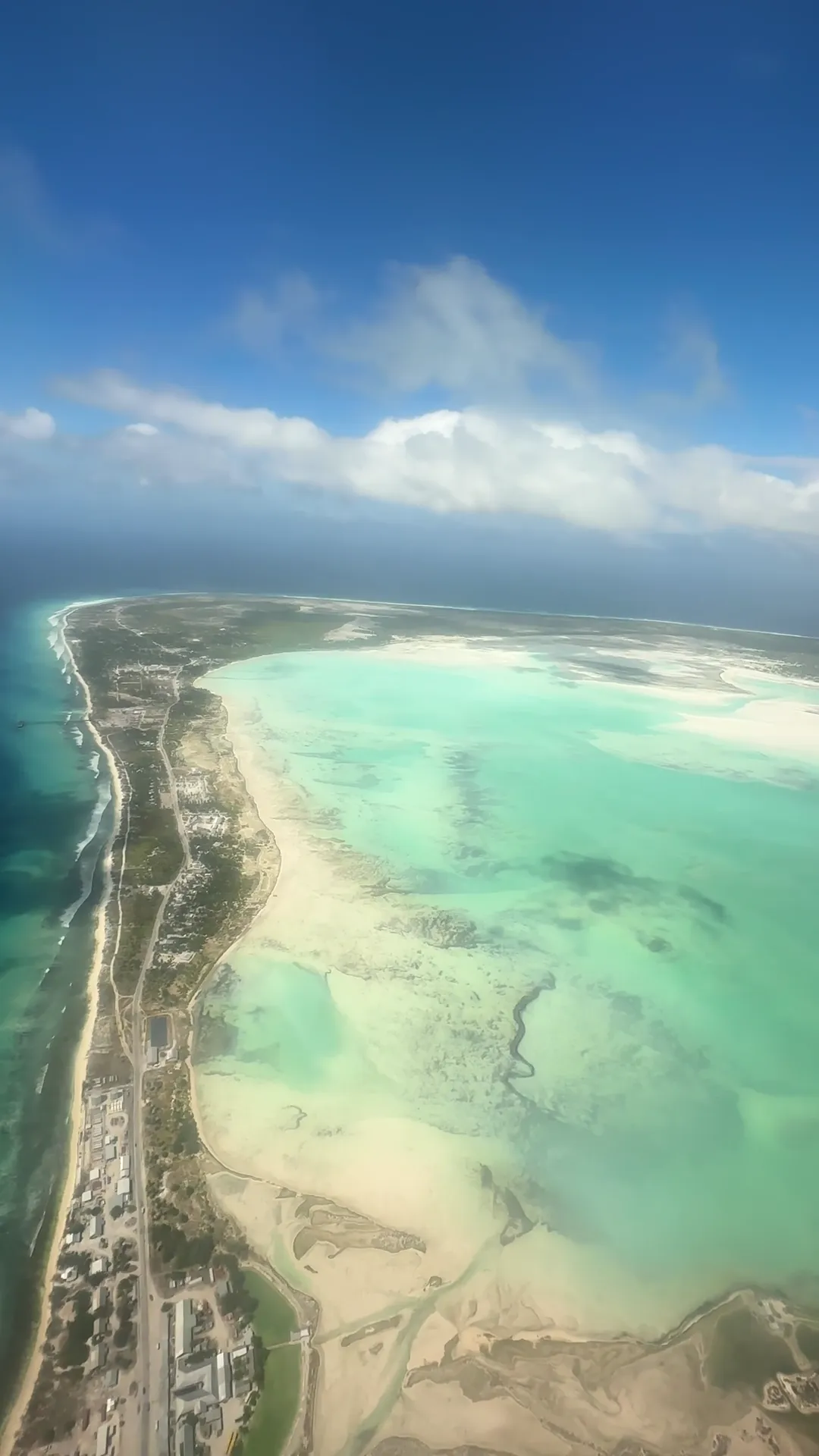 View from an aircraft window of the narrow strip of the London settlement between the dark blue ocean and a vast, shallow turquoise lagoon, clouds above, Christmas Island (Kiritimati), Kiribati