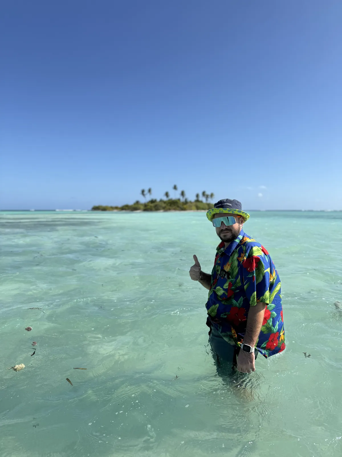 A person in a bright Hawaiian shirt, a sun hat and sports sunglasses standing waist-deep in a shallow turquoise lagoon and giving a thumbs-up; on the horizon a small palm-covered islet under a clear sky, Kiribati