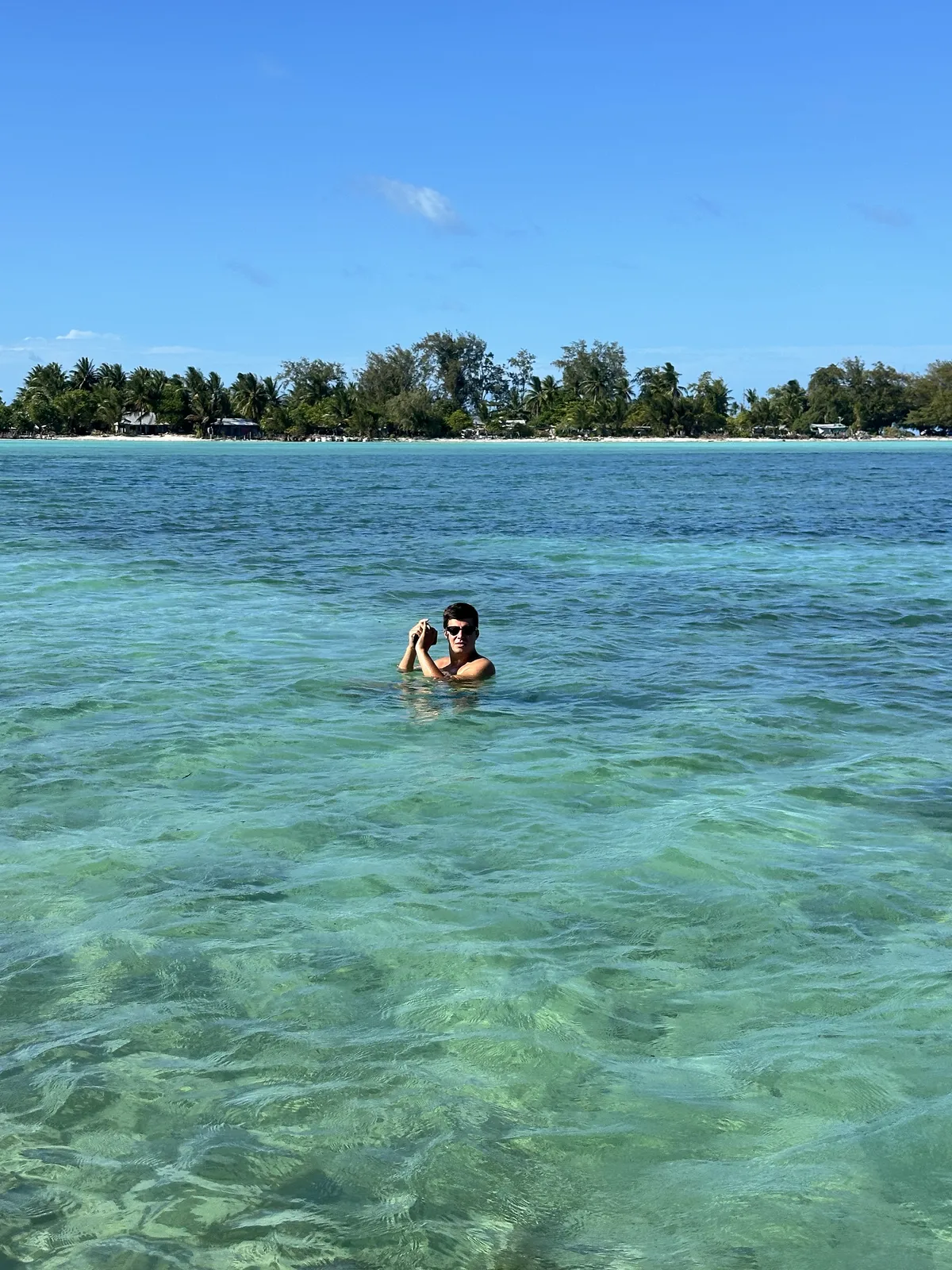 Timofei in sunglasses swimming in the clear turquoise lagoon of Tarawa, Kiribati, holding a camera; behind him a shore with palm trees and houses