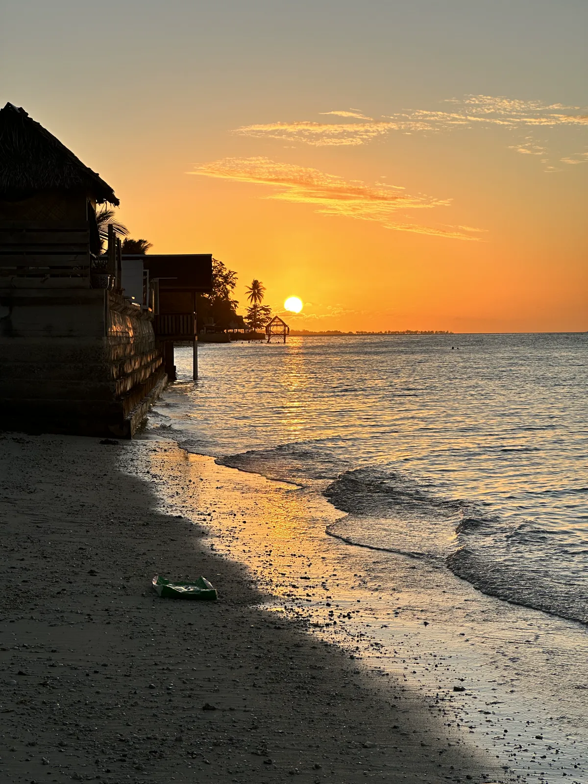 Sunset over the lagoon: a sandy beach, on the left the silhouette of a thatched bungalow on stilts, palm trees and the sun on the horizon, Tarawa (South Tarawa), Kiribati