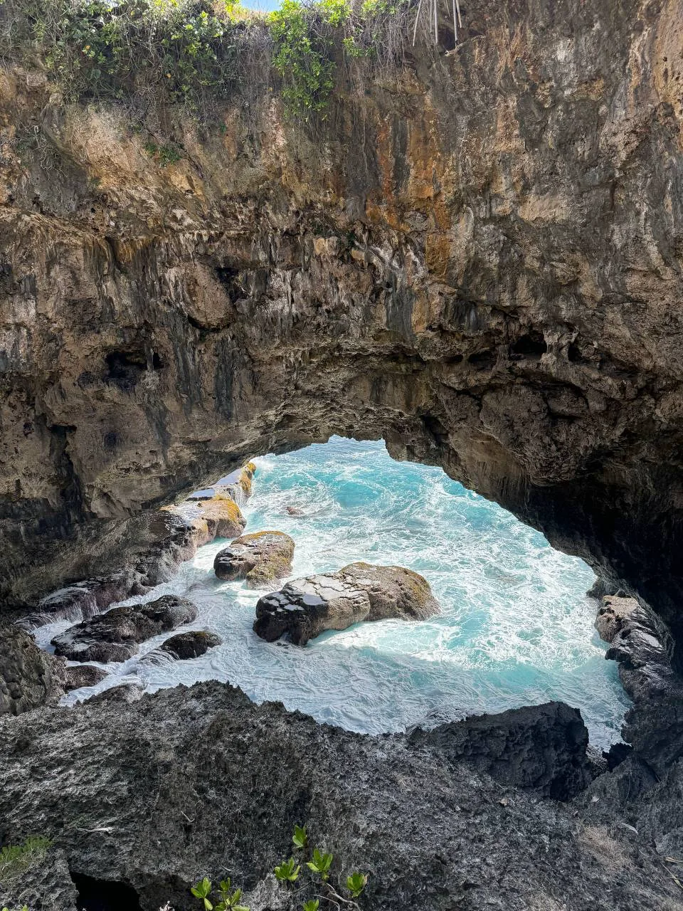 View from inside a natural stone arch: turquoise surf swirls around boulders under an overhanging limestone vault with greenery, Tonga