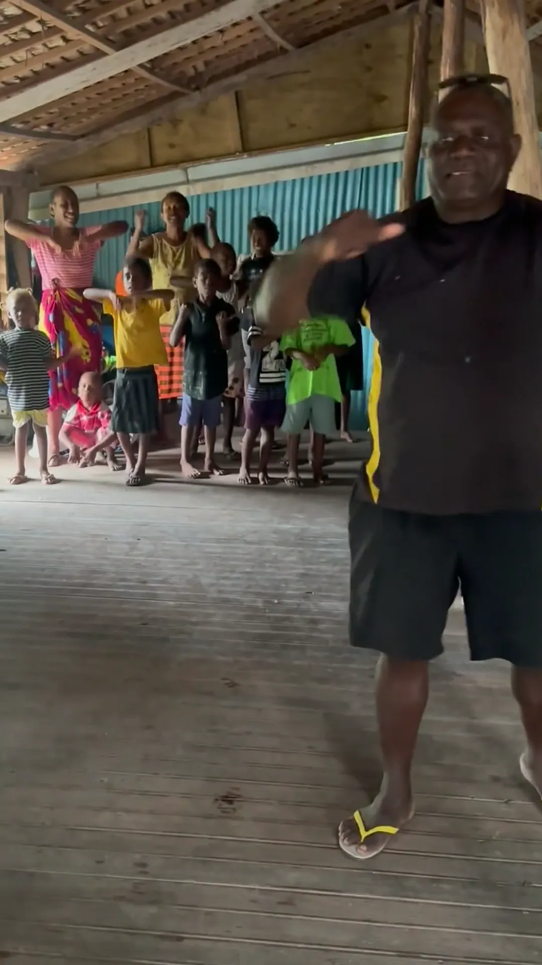 Children singing in a village hall under a palm-thatched roof on Lelepa Island, Vanuatu; a man in a dark shirt leads the singing